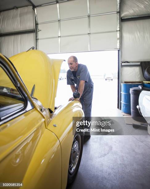 a mechanic looking under the bonnet of a bright yellow morris minor english classic car. - oldtimer werkstatt stock-fotos und bilder