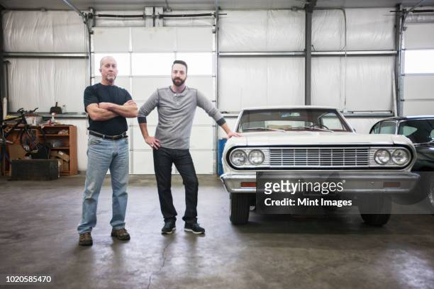 a portrait of a caucasian senior car mechanic and his son in their classic car repair shop. - old car garage stock pictures, royalty-free photos & images