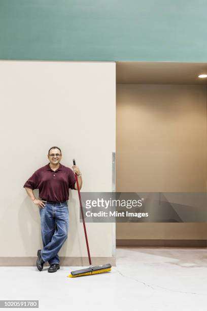 an hispanic workman standing in a large interior space with a broom. - bidello foto e immagini stock