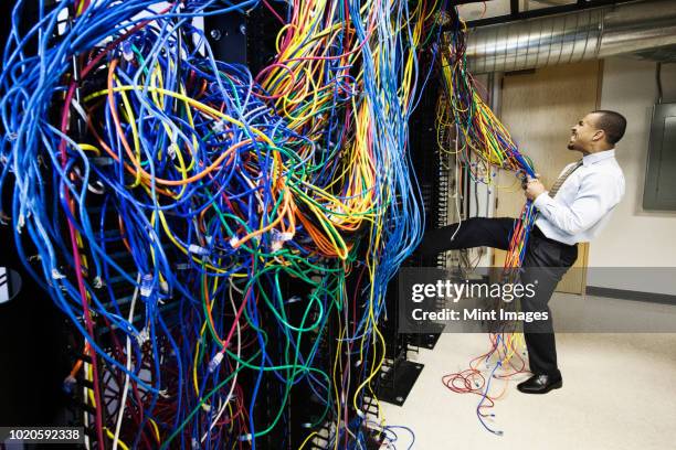 a black male technician pulling on a tangled mess of cat 5 cables in a computer server room. - network failure stock pictures, royalty-free photos & images