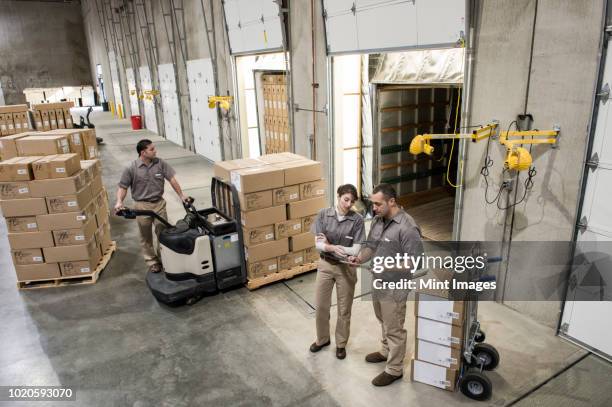 a team of three mixed race uniformed warehouse workers loading boxed products into the back of a truck in a distribution warehouse. - pallet industrial equipment fotografías e imágenes de stock