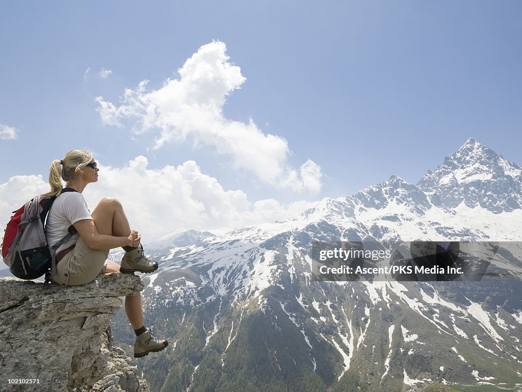 Hiker sits on rock shelf high above valley, mtns