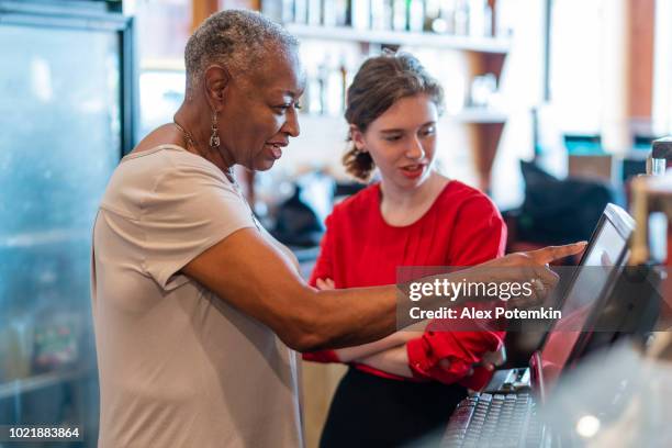 der aktive senior, 77 jahre alt, afroamerikanischen geschäftsfrau, unternehmer, die lehre des neuen mitarbeiters, der 18 jahre alte kaukasische weißes mädchen, wie mithilfe der edv kasse im kleinen lokalen restaurant. - black woman on cash register stock-fotos und bilder