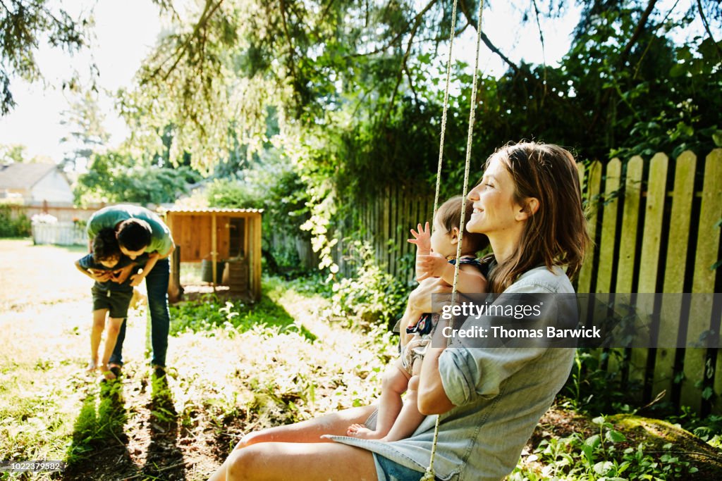 Mother holding infant daughter on swing while father plays with son in backyard