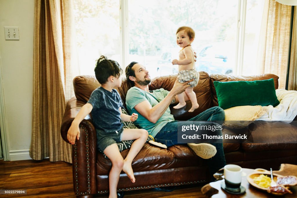 Smiling father holding up infant daughter while hanging out with children in living room