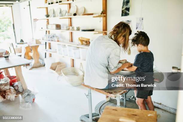 Young son helping mother make mug at potters wheel in studio in garage