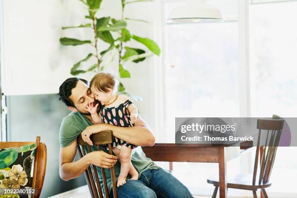 smiling father holding and kissing infant daughter in dining room in home - genderblend stock-fotos und bilder