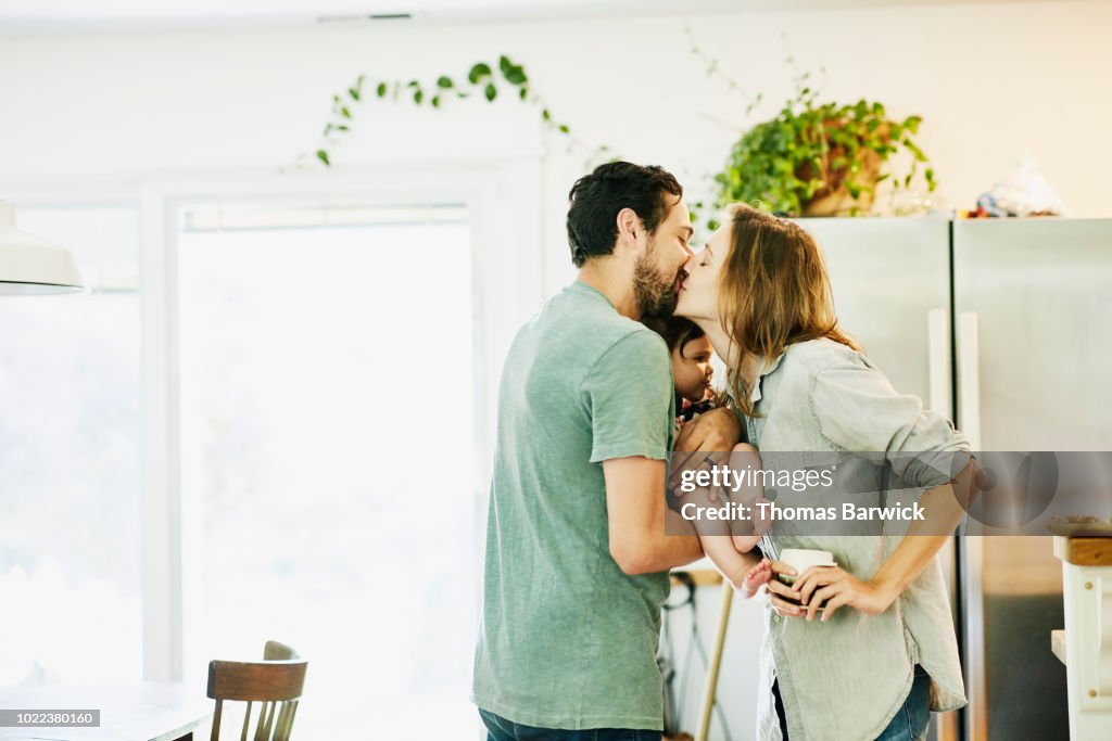 Mother and father kissing while holding infant daughter in kitchen