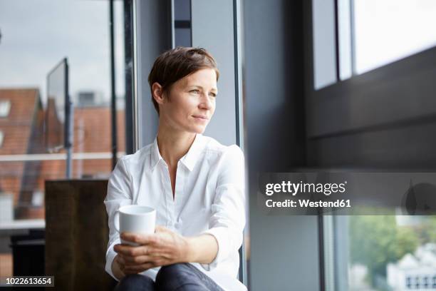 businesswoman sitting in office with cup of coffee looking out of window - coffee cup top view stockfoto's en -beelden