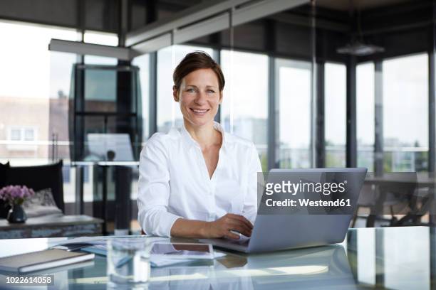 portrait of smiling businesswoman sitting at glass table in office with laptop - geschäftskleidung stock-fotos und bilder