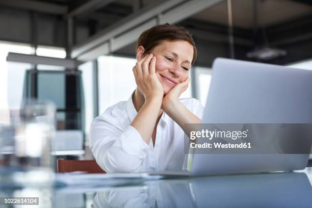 smiling businesswoman sitting at glass table in office looking at laptop - good news stock pictures, royalty-free photos & images