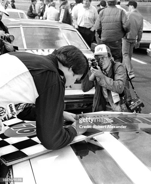 Driver Darrell Waltrip signs his autograph on a checkered flag as photographers take his photograph prior to the start of the 1980 Daytona 500 stock...