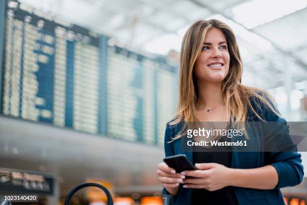 smiling young woman with cell phone at departure board looking around - airport departure board stock pictures, royalty-free photos & images