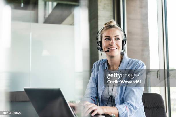 young businesswoman sitting at desk, making a call, using headset and laptop - headset imagens e fotografias de stock