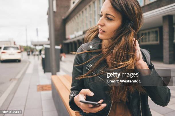 young woman with windswept hair holding cell phone in the city - chaqueta de cuero fotografías e imágenes de stock