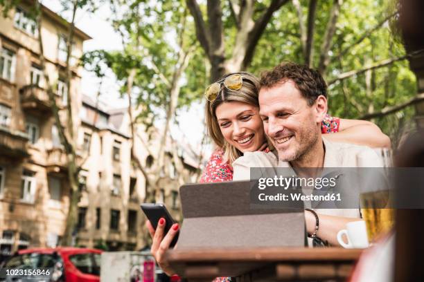 smiling couple sharing tablet and cell phone at an outdoor cafe - couple at table with ipad stockfoto's en -beelden