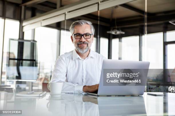 portrait of confident businessman with laptop at desk in office - punto di vista frontale foto e immagini stock