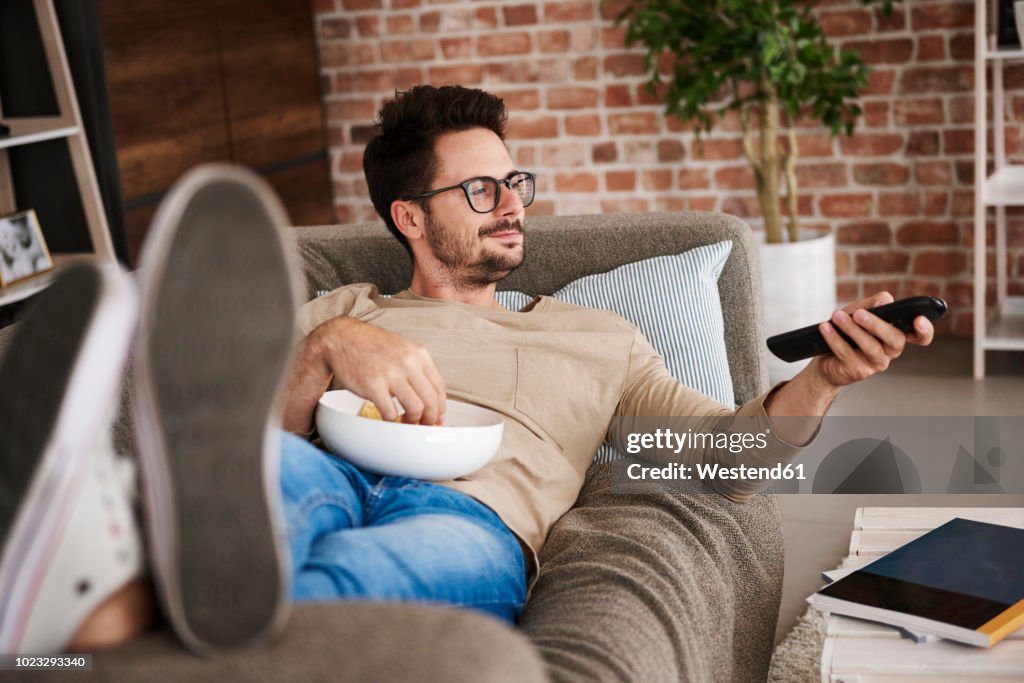 Content man lying on couch at home with bowl of potato chips watching TV