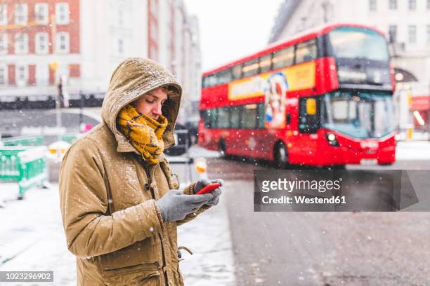 uk, london, young man standing next to the road in the city looking at cell phone - weather man stock pictures, royalty-free photos & images
