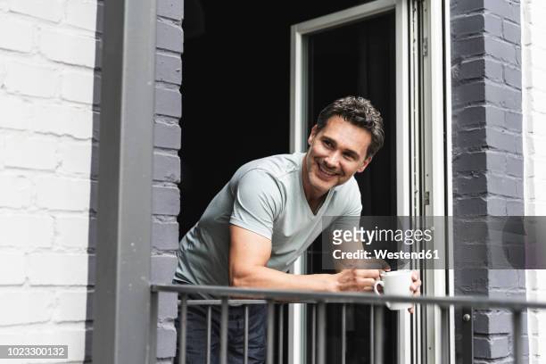 smiling man in pyjama at home with cup of coffee looking out of balcony door - leaning stockfoto's en -beelden