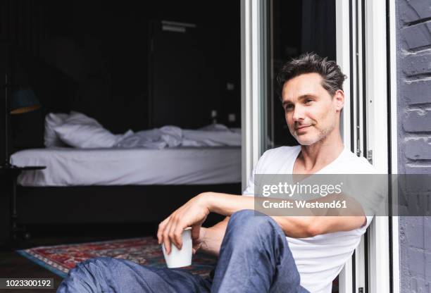 smiling man in pyjama at home with cup of coffee sitting at french window - early men stock-fotos und bilder