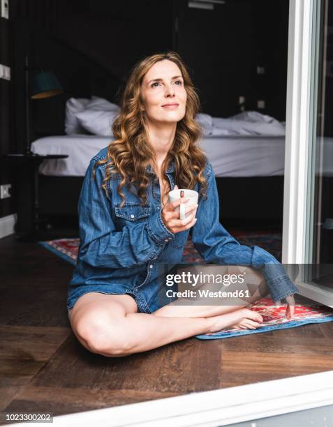 smiling woman in denim shirt at home with cup of coffee sitting at french window - denim shirt stock-fotos und bilder