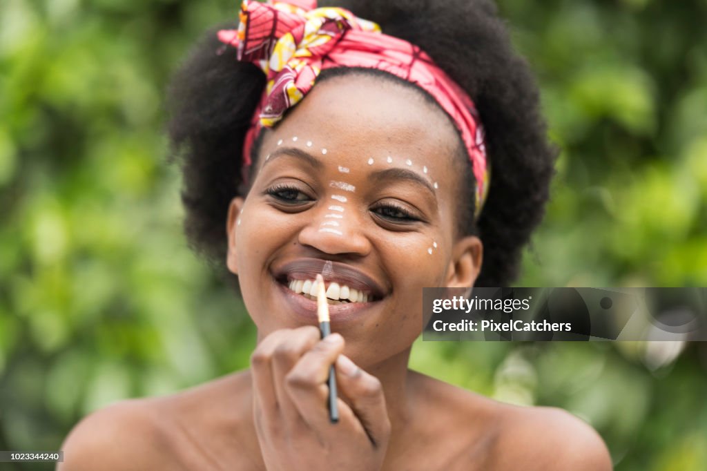 Young African woman smiles into mirror as she paint traditional make up patterns on her face