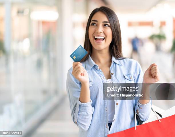 excited young woman looking away daydreaming while holding her debit card and shopping bags - credit card stock pictures, royalty-free photos & images