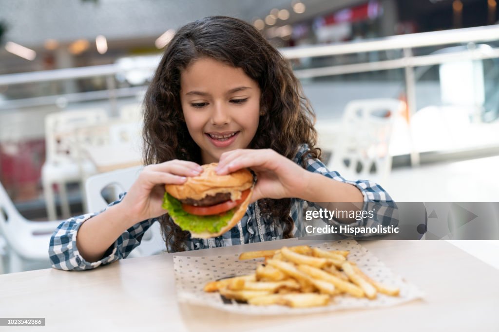 Wunderschönes kleines Mädchen genießen einen Burger im Foodcourt in einem Einkaufszentrum suchen sehr glücklich