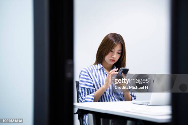 young woman texting with smart phone at laptop in office - premium access image only stock-fotos und bilder
