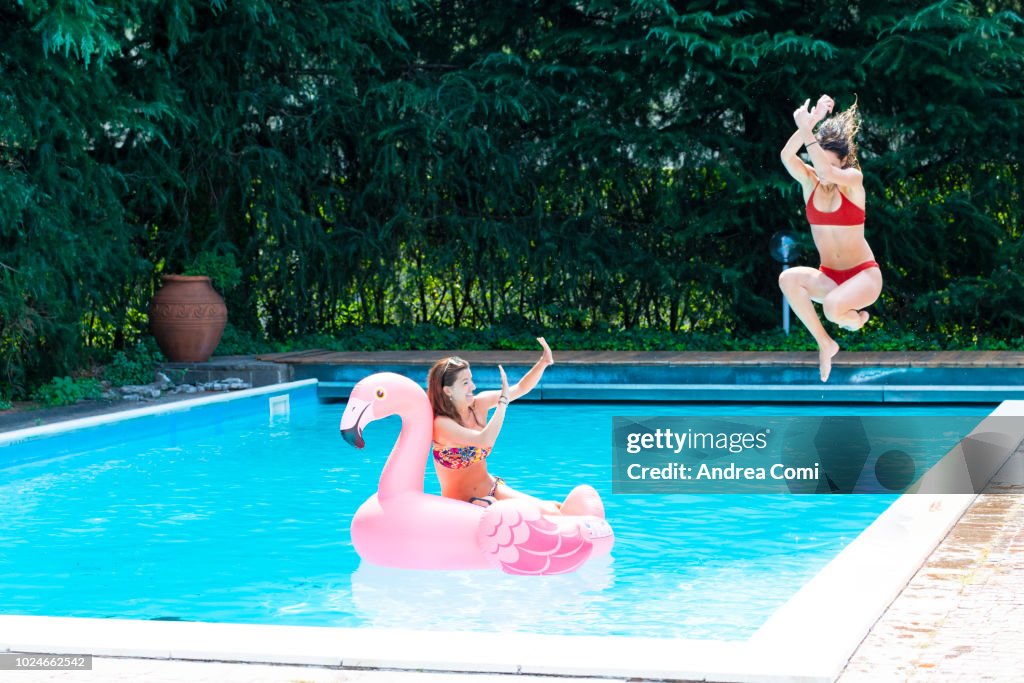 Young woman jumping into a swimming pool with a friend on a flamingo float