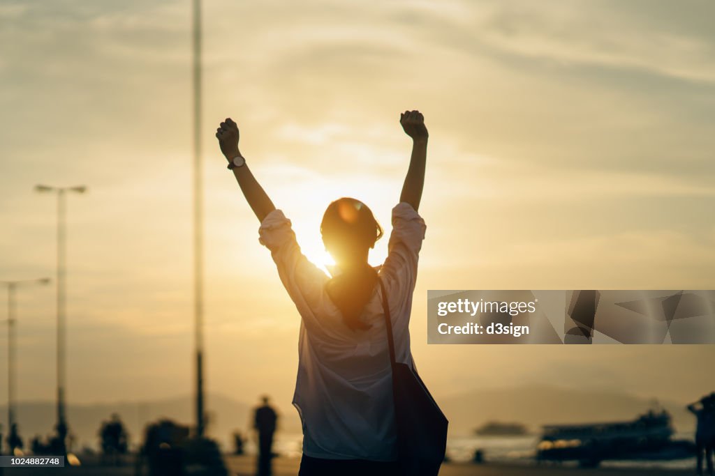 Young woman relaxing with hands in the air by the pier and enjoying the beautiful sunset and warmth of sunlight