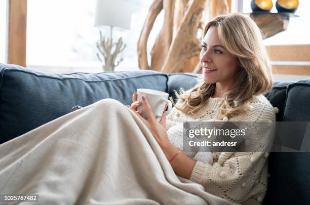 woman relaxing at home drinking a cup of coffee and lying on the sofa - winter sofa stock pictures, royalty-free photos & images