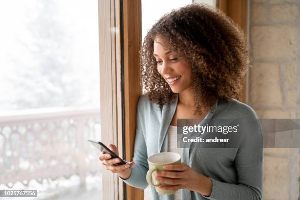 beautiful woman at home drinking a cup of coffee and texting on her phone - winter home stock pictures, royalty-free photos & images