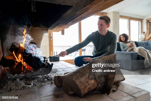 mann, die brennende maschinenbordbücher im kamin und entspannen sie mit seiner partnerin in ein winter-lodge - kamin konstruktion stock-fotos und bilder