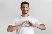 Portrait of smiling young man keeps hands on chest in heart shape sign, expresses sympathy. Kind hearted friendly nice guy wearing blank white t-shirt