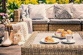 An outdoor wicker table and a sofa with cushions. Croissants for breakfast on a patio on a summer morning in an exclusive apartment with garden during vacation.