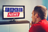 lies of tv propaganda mainstream media disinformation, A fake news report. viewer is watching TV and doesn't believe in fake news. man closes his eyes not to watch the lies on TV.