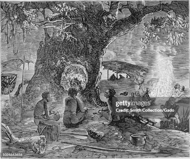 Black and white vintage print, depicting several Andamanese or Mincopies men, wearing loincloths, and seated on the ground in front of a 'tree oven'...