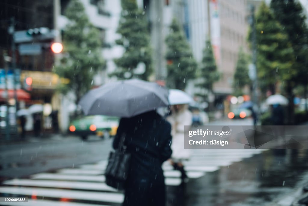 Defocused image of pedestrians carrying umbrella walking in downtown city street during rain