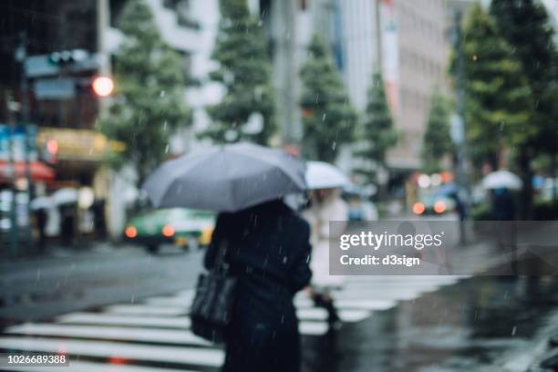 defocused image of pedestrians carrying umbrella walking in downtown city street during rain - rainy season stock pictures, royalty-free photos & images