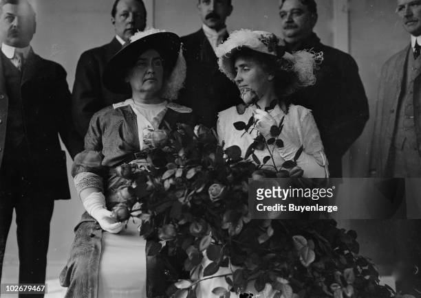 American teacher Anne Sullivan Macy and her former student, author and activist Helen Keller sit together while several dignitaries stand behind them...