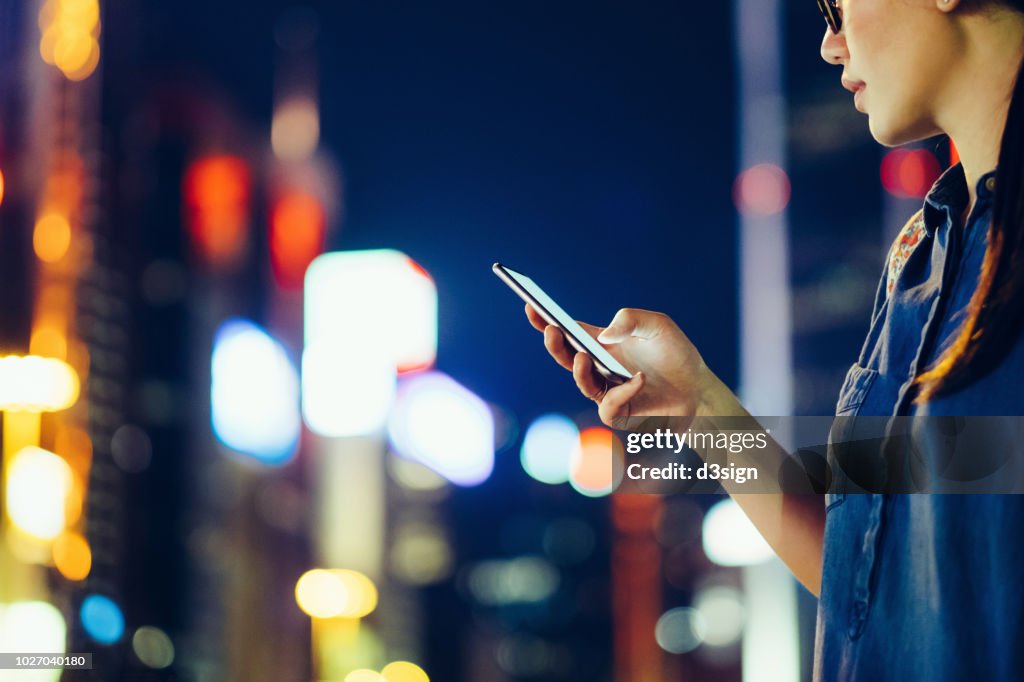 Cropped image of young woman using mobile phone outdoors at night, against city buildings with multi coloured neon lights