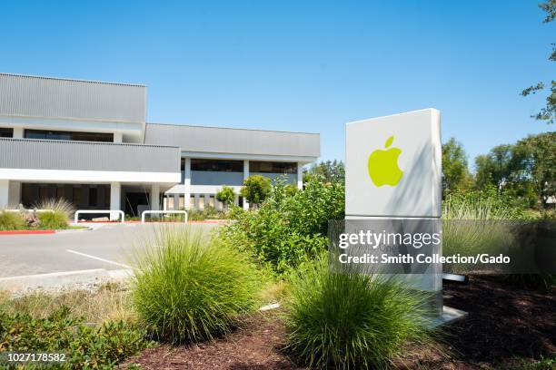 Sign with green colored logo amid landscaping, with facades of buildings in background, near the headquarters of Apple Computers in the Silicon...