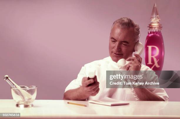 Pharmacist reads from the bottle of medicine, as he speaks on the phone, while filling out a prescription, 1988. A mortar and pestle, used for...