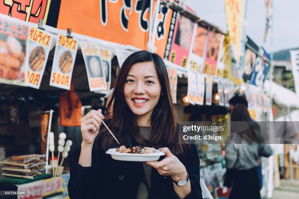 Joyful female tourist enjoying Japanese style snacks from local street vendor while travelling in Japan