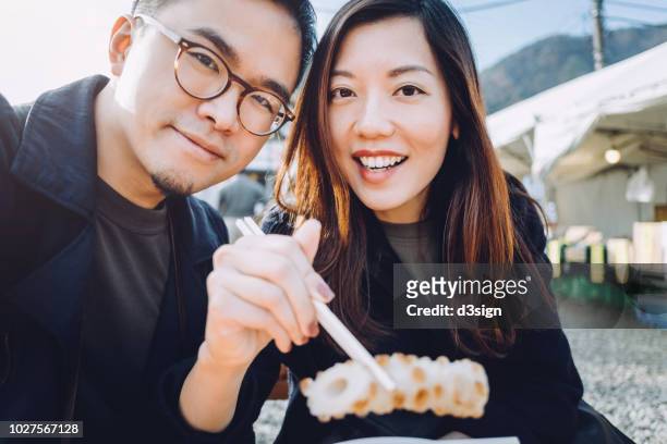 joyful asian couple tourist enjoying japanese snacks from local street vendor while travelling in japan - destination fashion 2016 stockfoto's en -beelden