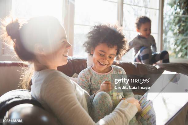 happy mother and daughter cuddling, reading book on sofa in sunny living room - london 2018 day 5 stockfoto's en -beelden