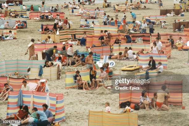 Crowd of holidaymakers use windbreaks to make the most of a British holiday on the beach at Perranporth, Cornwall circa 1970.