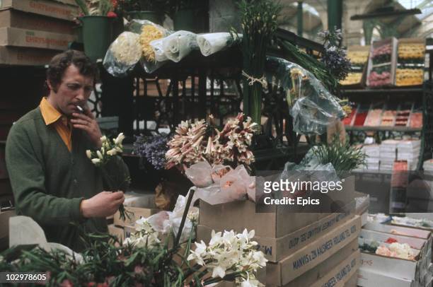 Flower seller takes a puff on a cigarette whilst working his stall in London's Covent Garden Market circa 1970.